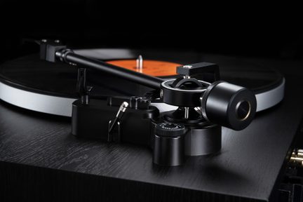 Närbild från sidan av en svart tonarm i metall med cylindriskt motvikt och silverfärgat ringformat pivot, monterad på svart träbas på en vinylspelare, svart vinylskiva med orange etikett i bakgrunden