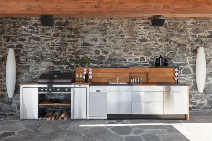 Buitenkeuken met houten werkblad, gootsteen, kookplaten, opbergruimte, tegen een stenen muur, twee opgehangen luidsprekers