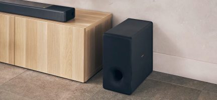 Ansicht eines hellen Holzmöbels mit einer schwarzen Soundbar darauf und einem schwarzen Subwoofer daneben auf dem Boden.