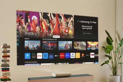 Væghængt skærm med Samsung TV Plus-interface, bogreol til venstre, grøn plante til højre, lavt møbel nedenunder