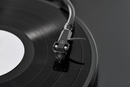 Detailní záběr na černé raménko gramofonu Audio-Technica na černé vinylové desce, šedé pozadí