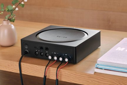 Čierny zosilňovač Sonos, pohľad zozadu, s audio konektormi a pripojenými káblami na drevenom stole