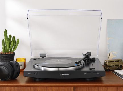 Sort vinylpladespiller i plast og metal, gennemsigtig hævet låg, skive på plade, tonearm, forfra set