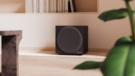 Mattschwarzer Subwoofer im würfelförmigen Gehäuse, frontale Ansicht leicht von oben, große zentrale runde Gitterabdeckung und kleine gelbe Kontrollleuchte unten links