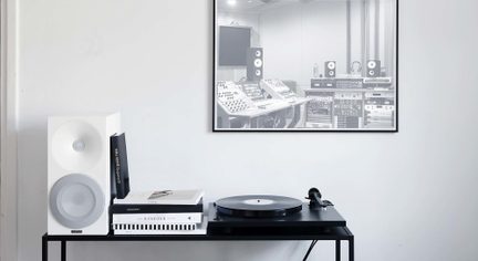 Zwarte platenspeler op een console met witte luidspreker en boeken, onder een fotolijst van een opnamestudio