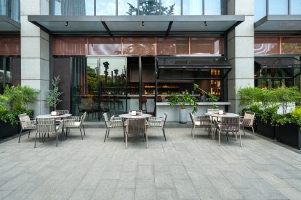 Terrasse mit runden Tischen, Stühlen mit durchbrochener Rückenlehne, grauem Plattenboden, Pflanzen in Töpfen, verglaster Fassade mit sichtbarer Bar