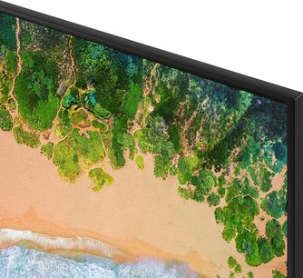 TV-skærm, der viser et luftfoto af en strand med turkisblåt vand og orangesand, omgivet af en frodig, grøn tropisk skov