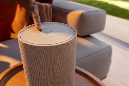 Zylindrischer, beigefarbener Bluetooth-Lautsprecher mit Touch-Bedienfeld oben, Hand drückt die Play-Taste