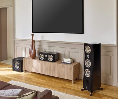 Stue, set forfra, sort vægmonteret tv, lavt møbel i lyst træ, sorte lakerede højttalere (højttalerkolonne, centerhøjttaler, subwoofer)