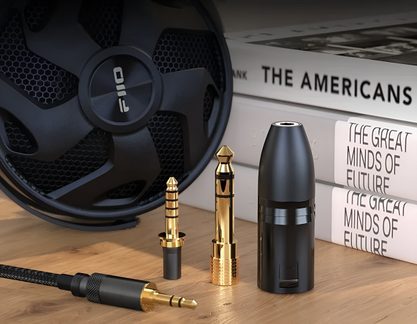Let svagt nedadgående nærbillede af et sæt gyldne jackstik i metal, ledsaget af en sort cylindrisk adapter i plast og et sort flettet lydkabel, arrangeret på et træbord med en sort højttaler og bøger i baggrunden