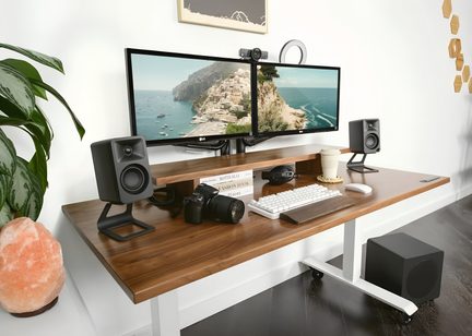 Arbejdsstation set fra trekvart vinkel, stort brunt skrivebord med hvidt metalstel udstyret med to sorte skærme på hylde, hvidt mekanisk tastatur med håndledsstøtte i træ, kompakte sorte højttalere, spejlreflekskamera, lys kop og diverse tilbehør