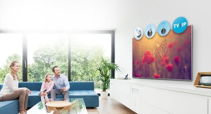 Família sentada em um sofá azul na sala de estar, assistindo a uma grande TV de parede exibindo uma imagem de flores vermelhas e uma borboleta