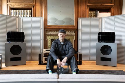 Grijze hi-fi-luidsprekers met zwarte hoorns in een woonkamer met houtafwerking, met een zittende man in het midden