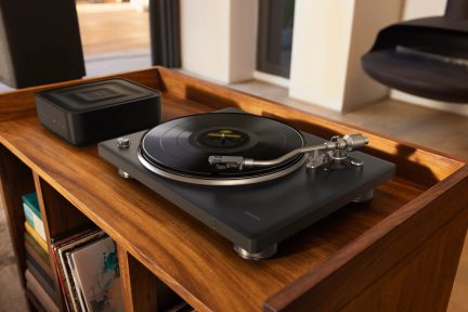 Тъмносив грамофон Denon с черна плоча и извито сребристо рамо, поставен върху дървен шкаф