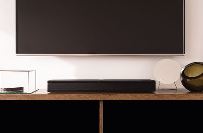 Sort Orbitsound ONE P70W soundbar på træmøbel, set forfra, med slukket tv og dekorative genstande