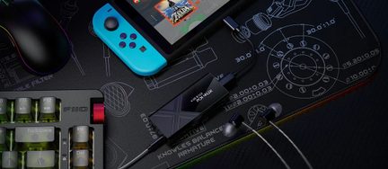 Bovenaanzicht van een set gamingaccessoires bestaande uit een zwart mechanisch toetsenbord van metaal en transparant plastic, een zwarte RGB-verlichte muis van plastic, een zwarte draagbare console met turquoise Joy-Con van plastic, een kleine glanzend zwarte USB-audio-omzetter van metaal met gevlochten kabel en zilverkleurige bedrade oordopjes van metaal, allemaal geplaatst op een zwart stoffen muismat bedrukt met technische schema’s.