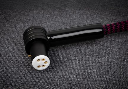 Vista ravvicinata dall’alto di un connettore angolato nero in plastica lucida con terminale cilindrico nero opaco e parte frontale bianca dotata di quattro pin metallici dorati, collegato a un cavo intrecciato nero e fucsia