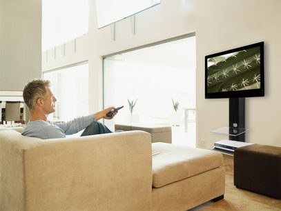En mand sidder på en beige sofa og peger med en fjernbetjening mod et sort TV-møbel med en fladskærm, der viser et mønster af stjerneblade