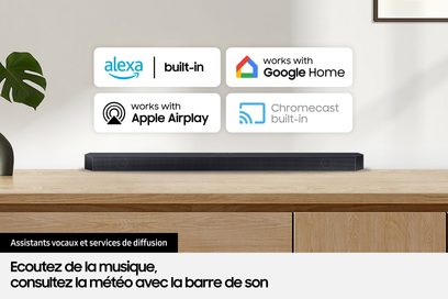 Samsung F-HW-Q995D-BF sort soundbar på lys træmøbel, grøn plante til venstre, fire logoer for stemmeassistenter ovenover