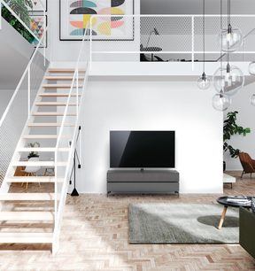 Lavt gråt antracitfarvet TV-møbel med glashylder, i moderne loftsmiljø med hvid trappe og glaspendellamper