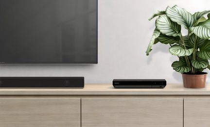 Rektangulær svart mediespiller sett forfra på et lyst tremøbel ved siden av en potteplante og en lydplanke