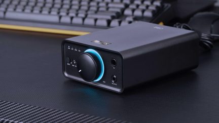 Matte black rectangular metal case, three-quarter front view, featuring a large volume knob encircled by a blue LED ring, two front-facing jack sockets, and a selection switch