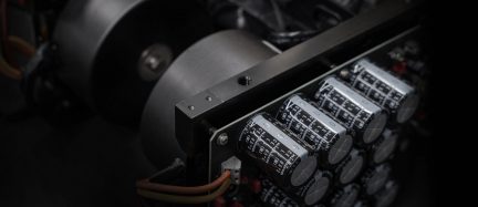 Reihe schwarzer zylindrischer Elektrolytkondensatoren mit Aluminium-Enden, montiert auf schwarzem Metallchassis, schräge Nahaufnahme, die im Hintergrund auch einen grauen Ringkerntransformator zeigt