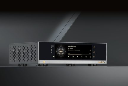 Lecteur réseau hi‑fi Matrix Audio en aluminium argenté, façade avant noire vitrée vue trois‑quarts, écran couleur central affichant la lecture, bouton doré à gauche, logo doré à droite, côtés perforés de grilles circulaires et quatre pieds cylindriques noirs