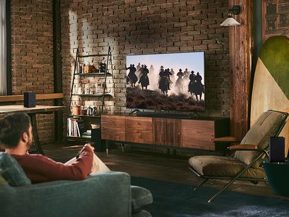 Fladskærms-tv der viser cowboys til hest i en stue med murstensvæg, reol, læderbrun sofa og lænestol