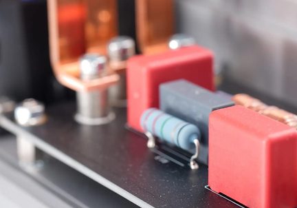 Zbliżenie ukośne czarnej płytki drukowanej z prostokątnymi czerwonymi obudowami z tworzywa sztucznego, cylindrycznym jasnoniebieskim rezystorem z kolorowymi pierścieniami oraz zakrzywionymi prętami z błyszczącej miedzi.