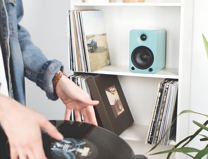 Boxă compactă turcoaz din plastic lucios, văzută din față, cu tweeter și woofer negre și două orificii frontale, așezată pe un raft alb printre discuri de vinil