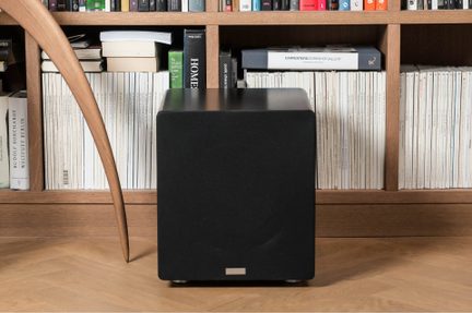 Sort, kubeformet subwoofer med stof-front, placeret på gulvet foran en træbogreol fyldt med bøger