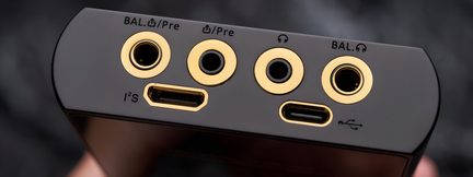 Gros plan de la tranche supérieure d’un boîtier audio noir mat en métal, alignant quatre prises jack circulaires dorées, un port I²S rectangulaire doré et un port USB-C