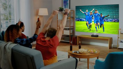 Tre personer sidder på en sofa og ser på en skærm, der viser fodboldspillere i blåt, som fejrer på en bane.