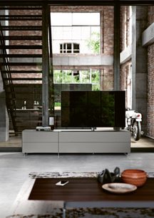 Lavt gråt TV-møbel med to skuffer, en TV-skærm, i en loftsleilighet med mursteinsvegger og store vinduer, interiørvisning