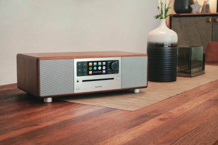 Компактна кафява и сива hi-fi уредба с LCD екран, бутони и високоговорители, поставена върху дървена маса