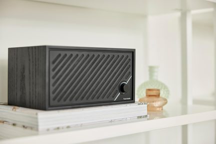Equipo de audio rectangular en madera negra, frontal con rejilla diagonal y botón giratorio a la derecha, vista desde tres cuartos izquierda