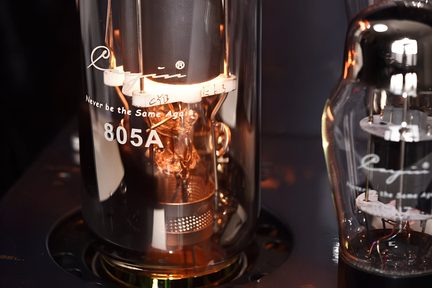 Let nærbillede let forfra af en cylindrisk elektronisk lampe i transparent glas, med sort, skinnende metalbase fastgjort i et mørkt chassis, indvendig struktur i glødende kobberfarvet metal, hvid inskription 805A på siden