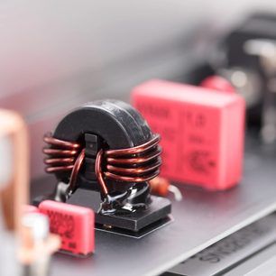 Detailní šikmý pohled na vícezásuvkovou přepěťovou ochranu Inakustik AC-3200 Filter Analog | SCHUKO, s černým feritovým toroidním jádrem obtočeným smaltovanými měděnými dráty, doplněnou růžovými obdélníkovými kondenzátory