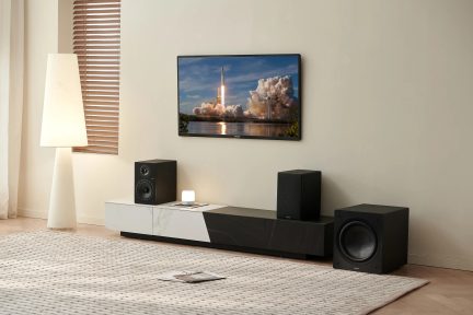 Svarte bokhyllehøyttalere på en hvit og svart TV-benk og en subwoofer på gulvet i en stue med veggmontert TV