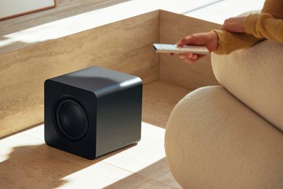 Černý matný kovový krychlový subwoofer se zaoblenými hranami, kruhový boční reproduktor na levé straně, pohled z levého předního tříčtvrtečního úhlu