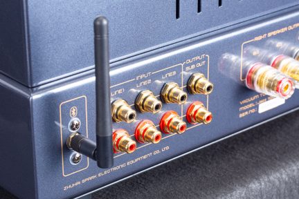 Detailní pohled ze tří čtvrtin zezadu na kovový audio zesilovač v antracitově modré barvě s matným povrchem, vybavený černou Bluetooth anténou a více RCA konektory pozlacenými s červeným lemováním.