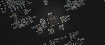 Scheda elettronica in fibra di vetro nero opaco, vista macro dall’alto, centrata su un circuito integrato quadrato a piedini sottili, circondato da resistenze e condensatori SMD serigrafati in bianco