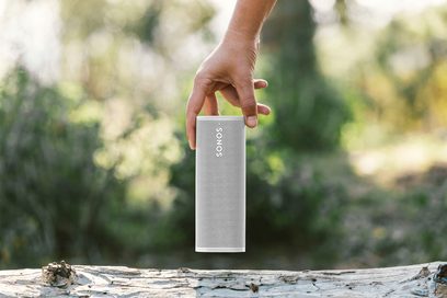 Bílý bezdrátový reproduktor Sonos Roam SL umístěný na kmeni stromu s rozmazanou vegetací v pozadí