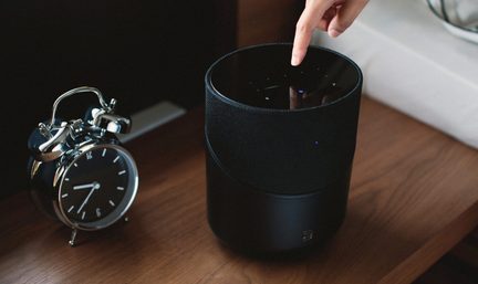 Hlavní ukazující na černou válcovou chytrou reproduktorovou jednotku vedle černého budíku na dřevěném povrchu