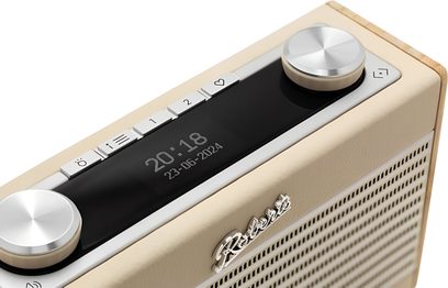 Radio til bordet Roberts Rambler Uno i beige med kornet kunstlæder og træsider, set fra trekvart højde, der viser den perforerede front, kromlogo, centralt sort blankt display med ur, fem lysegrå rektangulære knapper og to brede håndtag i børstet aluminium
