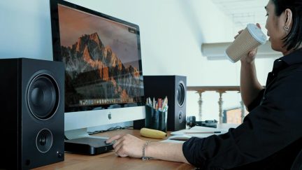Dataskjerm med bilde av fjell, svarte høyttalere, hånd som holder kopp, tastatur, mus, penner, banan