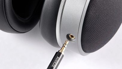 Close-up side view of the Audeze LCD-S20 closed-back wired hi-fi headphones in metallic gray aluminum, featuring black faux leather ear cushions, black fabric grille, and a gold-plated 3.5 mm jack being inserted