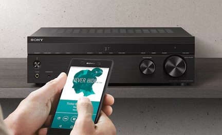 Juodas stiprintuvas su apvaliais mygtukais, juodas išmanusis telefonas, kuriame rodoma muzikos programa, rankos laikančios išmanųjį telefoną.