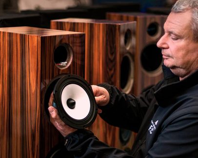 Enceinte colonne en placage bois brun veiné avec évent circulaire supérieur et logement vide, vue de trois-quarts, un homme tenant un haut-parleur détachable à membrane blanche et cadre noir devant l’ouverture